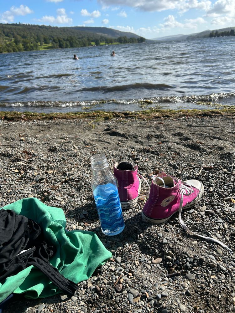 Still life: abandoned pink Converse, water bottle towel, on the lake shore with two distant figures far out in the lake 