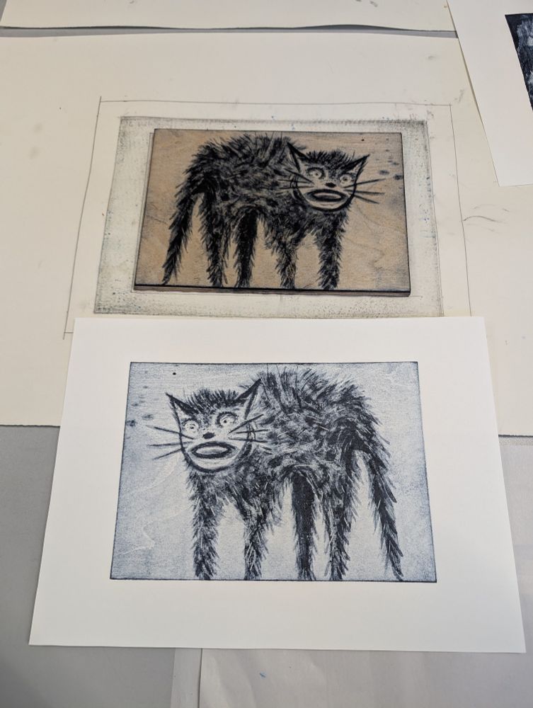 At the tip of the photo is a plywood block with a cat drawn on it in lithographic ink. Beneath it is a piece of paper with the reverse image printed on it. Both images are black and white.
