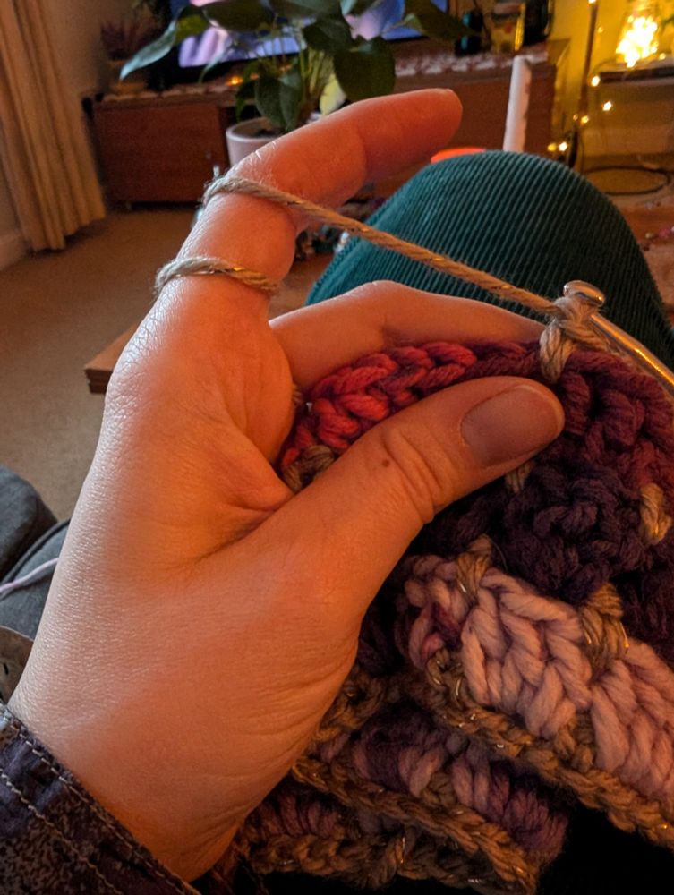 A hand with yarn wrapped around the forefinger holds the very beginning of a purple and pink crocheted blanket.