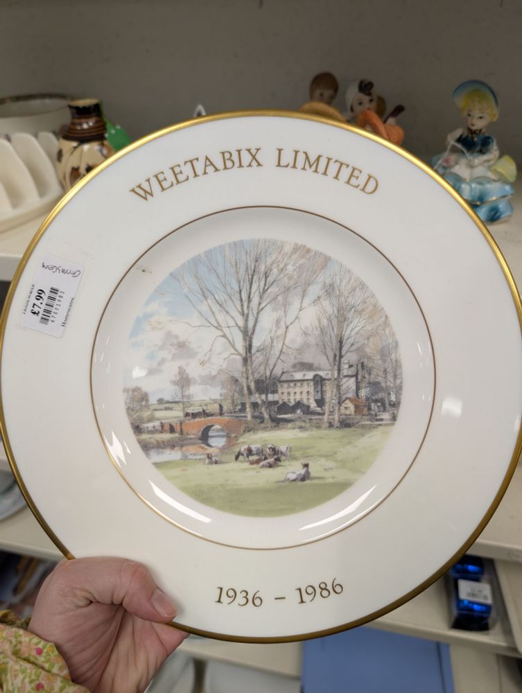 A commemorative plate for Weetabix with gold edging and a pastoral image of cows, a bridge and a factory in the background. Gold letters spell out "Weetabix Limited 1936-1986"