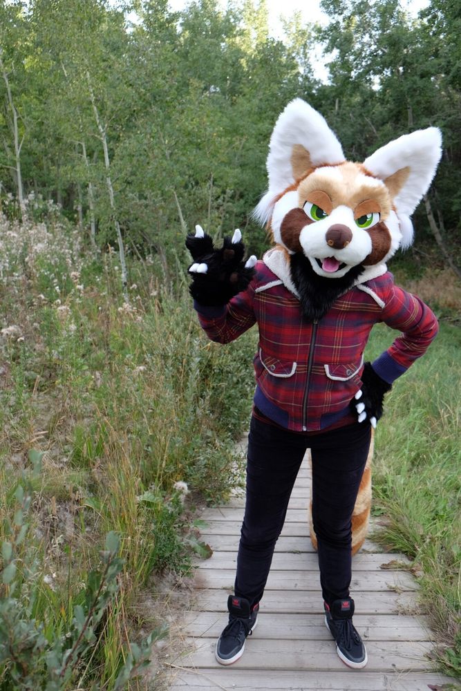 A red panda fursuit with green eyes, looking toward the viewer with angry eyelids and making a 'rock on' sign with their paw, wearing a red plaid jacket and standing on a boardwalk in a forest valley