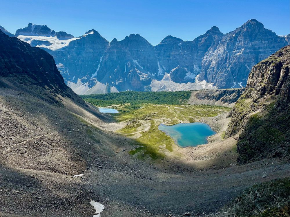 A row of jagged peaks are in the background with a glacier on the left side. Two small blue lakes sit in meadows in the center flanked by cliff walls on both sides. The trail is visible winding from the right-hand lake up the rocky slope on the left.