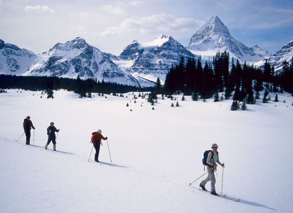 Four cross-country skiers glide across flat snow with their shadows before them. The background fills with jagged peaks including the tallest on the right shaped like a pyramid.