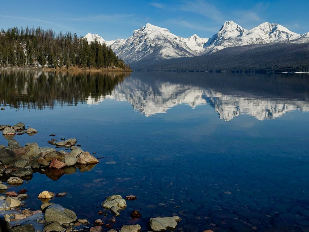 A glassy lake reflects snow-covered peaks and forested hillsides. Colorful shoreline rocks are visible on the bottom left. 