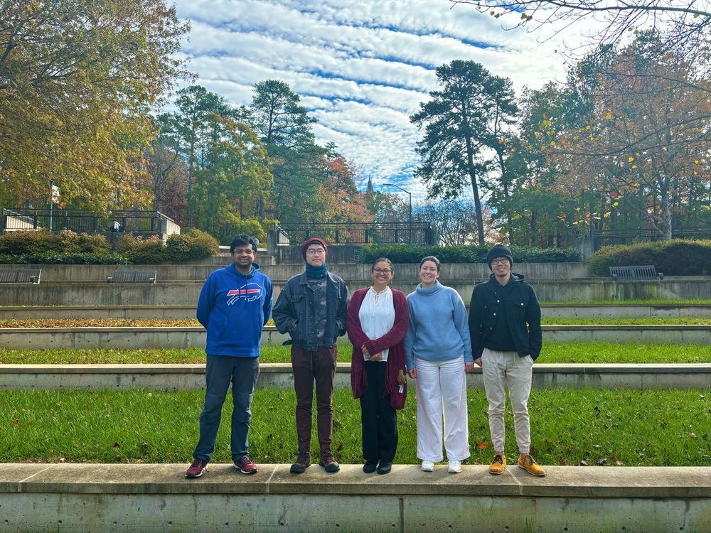 Five members of the Mazo-Vargas laboratory are standing at the Duke University campus, with tiered grassy steps and autumn trees, under a sky with scattered clouds. 