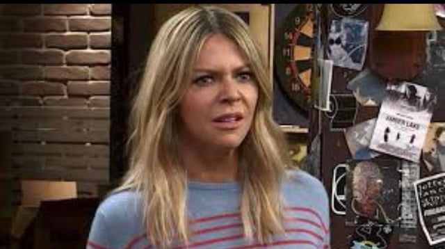 Sweet Dee, a blonde woman stands in a dimly lit bar, staring ahead with a confused, slightly alarmed expression. Her posture and narrowed eyes give her a subtle, birdlike alertness, as if she’s noticed something shiny or threatening offscreen. Behind her, a cluttered bulletin board and dartboard add to the chaotic atmosphere.