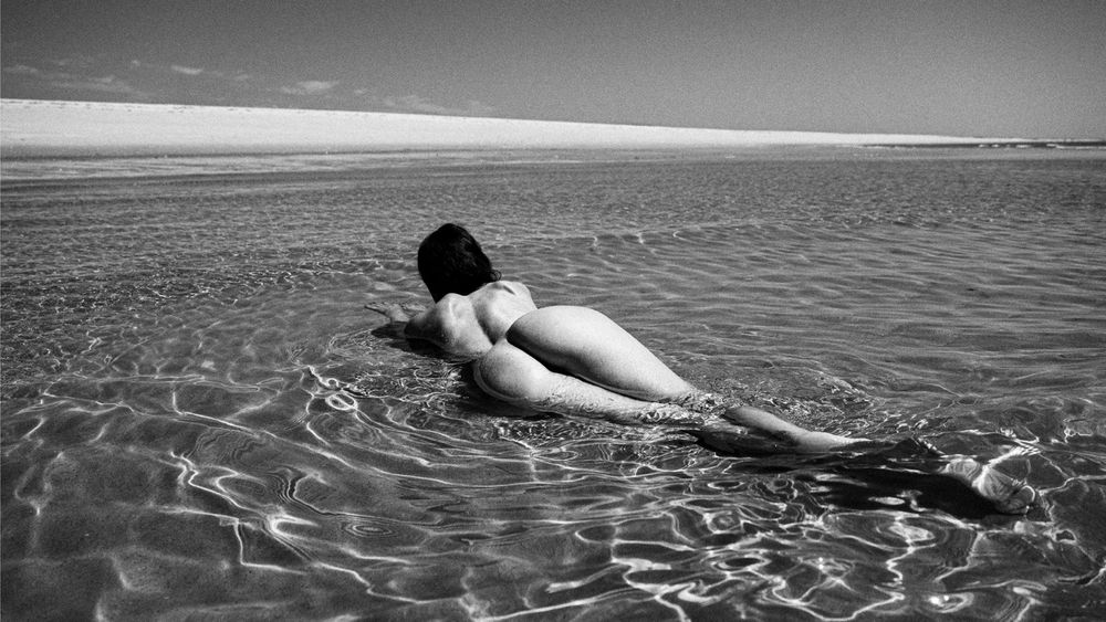 Eine Frau liegt nackt mit dem Bauch im flachen, klaren Wasser eines weiten Strandes, ihr Körper von der Sonne beschienen, während sich Lichtreflexe auf der Wasseroberfläche spiegeln. Der Horizont ist weit und leer, die Atmosphäre ruhig und zeitlos.