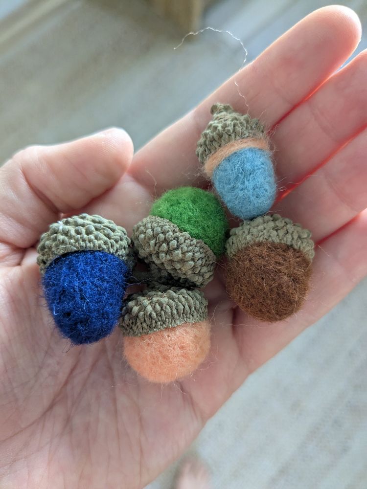 A white person's palm displaying several small felted acorns glued into real acorn caps