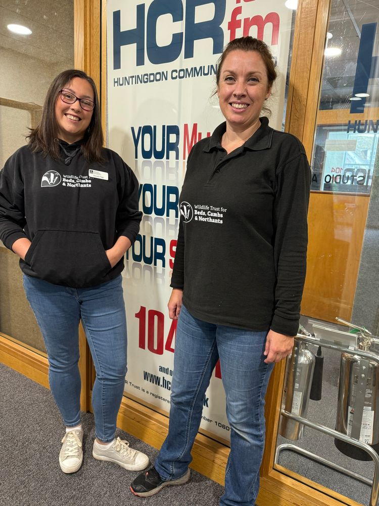 Danielle and Alison, in black wildlife Trust branded tops and jeans, stand in front of a wall branded with HCR104fm