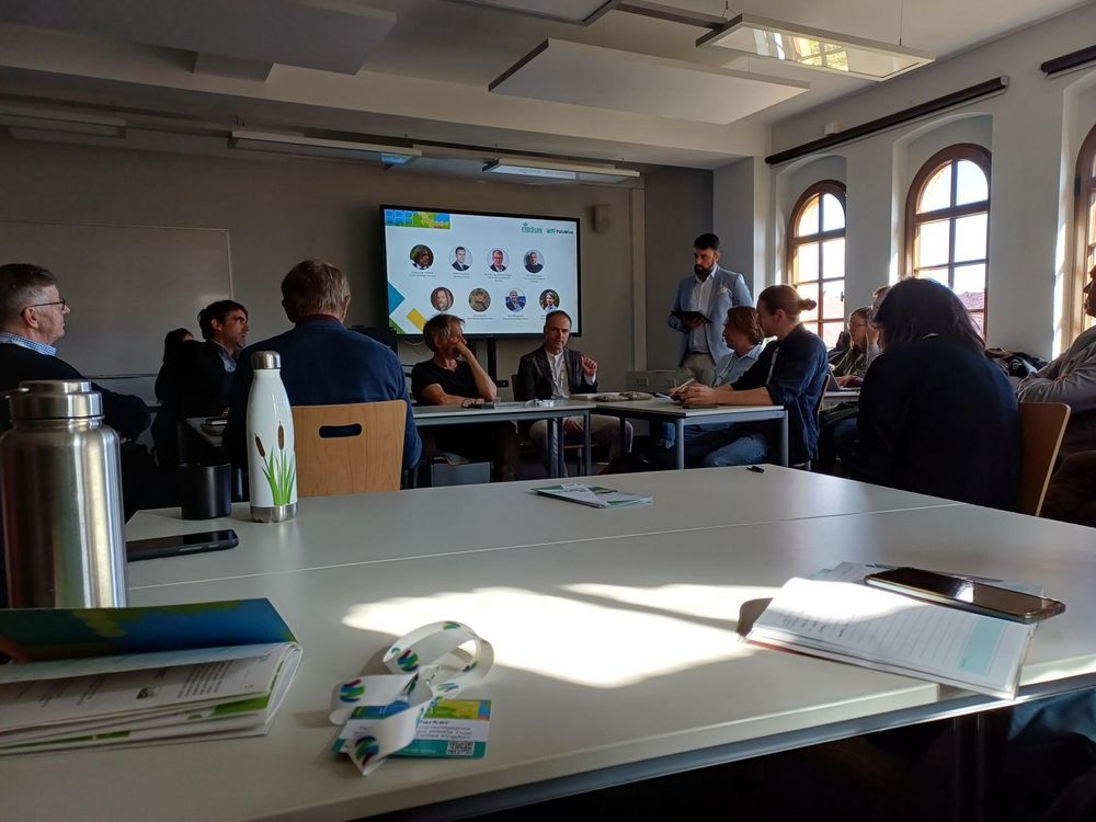 Conference attendees sit at tables in a square around a room in discussion