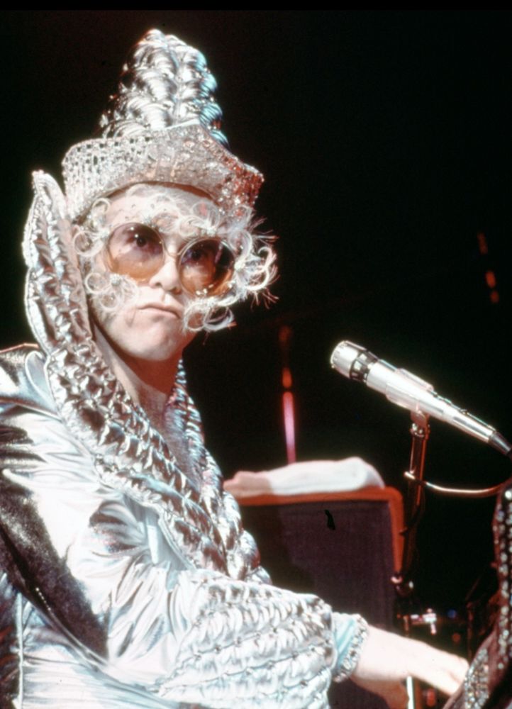 young elton john in a silver outfit and big feathery glasses