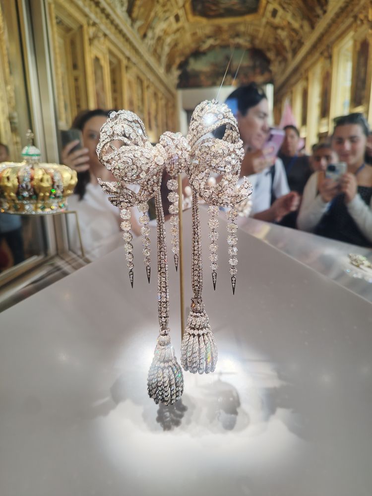 foto de um broche de napoleão. é um laço feito em metal prateado, com pendente também em metal, todo cravado de brilhantes