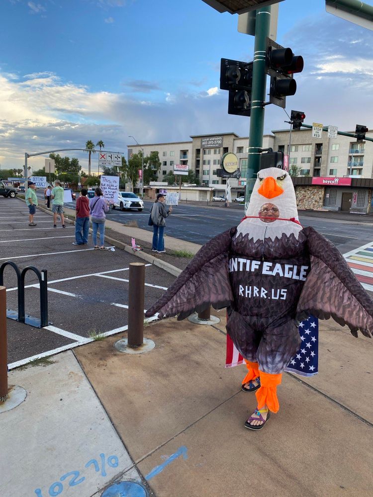 An inflatable eagle with “Antifeagle” and “RhRR.us” written in white stands near a street with other protesters in the background. 