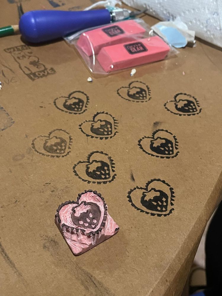 Image showcases a carved out eraser used as a stamp. The stamp is a strawberry inside an uneven trim heart. The background is brown craft paper, which has  impressions of the stamp with black ink. The materials used to craft are also shown in the back.  