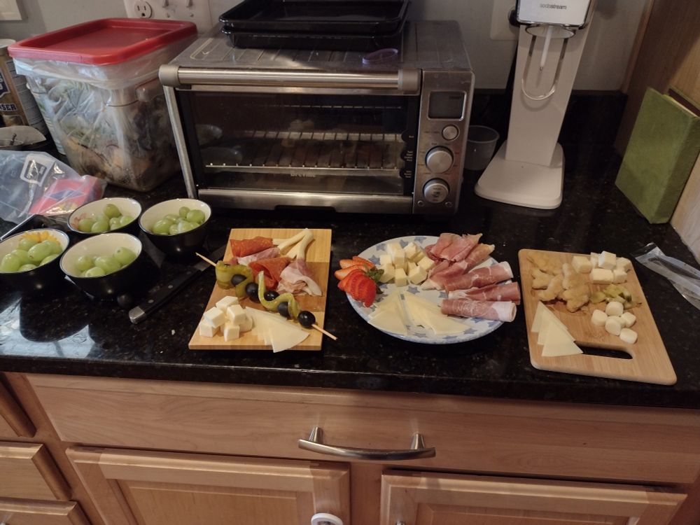 A dark countertop with three different cheese and charcuterie plates.  On the far left are four small bowls of grapes.  The back of the counter area has other kitchen appliances.