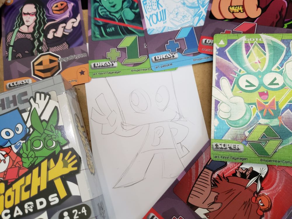 a photo of a small pencil sketch of Paper from Gotcha Cards, surrounded by various promotional cards from the Kickstarter campaign and a pack of the card game itself