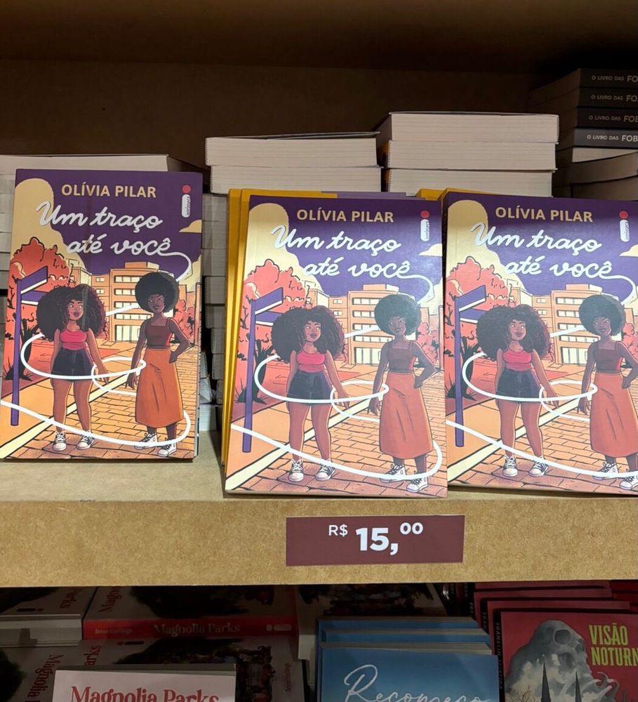 estante com livros Um traço até você e destaque para preço R$15

Capa de Um traço até você. Ilustração de duas garotas negras no centro, a da esquerda tem o cabelo volumoso e crespo, ela tem lábios grossos e está olhando para a garota ao seu lado, sua pele é marrom médio – ela também veste um short preto e uma camisa rosa. A garota da direita tem o cabelo crespo em black power, usa uma saia e cropped em tons terrosos, sua pele é marrom escura. Elas estão com as mãos erguidas quase se tocando. O título da autora está em amarelo, o mesmo tom dos prédios ao fundo. Título do livro em branco, com um traço que puxa ao redor das personagens. No fundo da ilustração, prédios em tons de laranja e amarelo, e o céu tem o tom roxo. Logotipo da editora Intrínseca no canto superior direito.