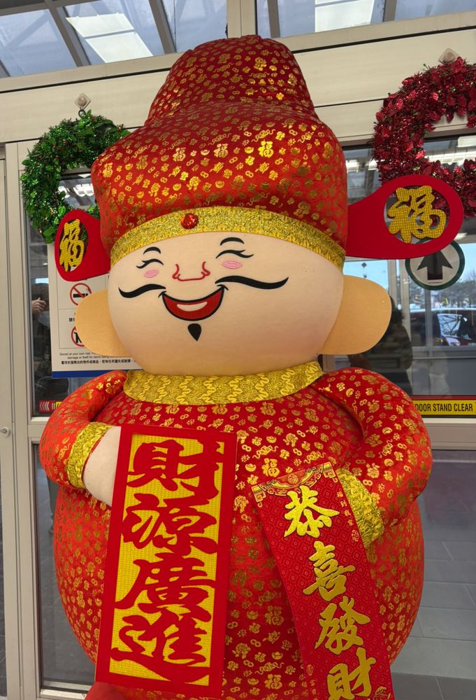 A large statue of a cartoon-like man dressed in red and gold holding good luck signs written in Chinese.