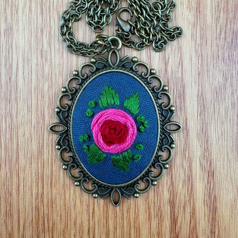 An embroidered rose surrounded by four leaves on a blue fabric, set into a brass-coloured necklace.