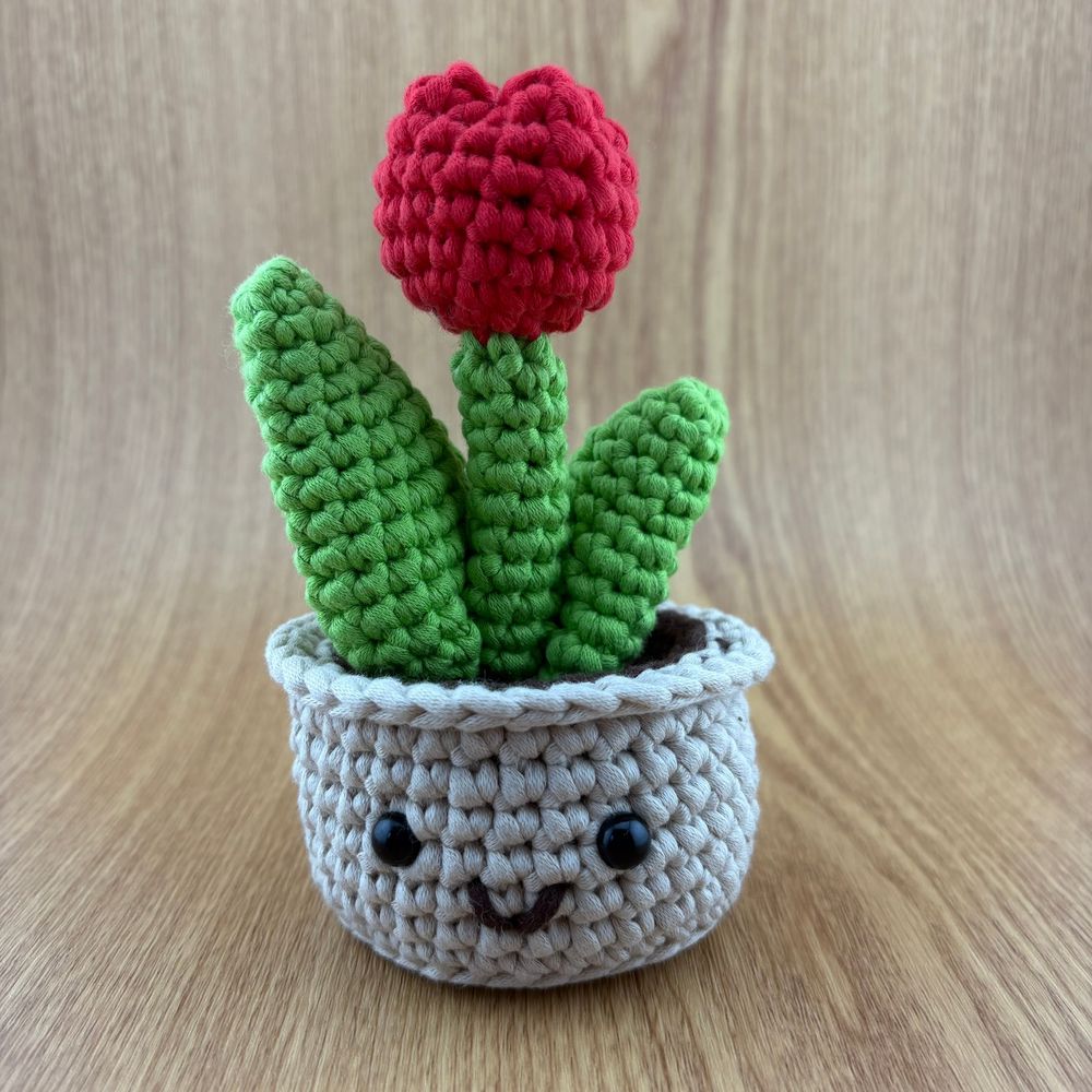 A picture of a red crocheted tulip with two green leaves in a light brown crocheted pot. The pot has a smiling face.