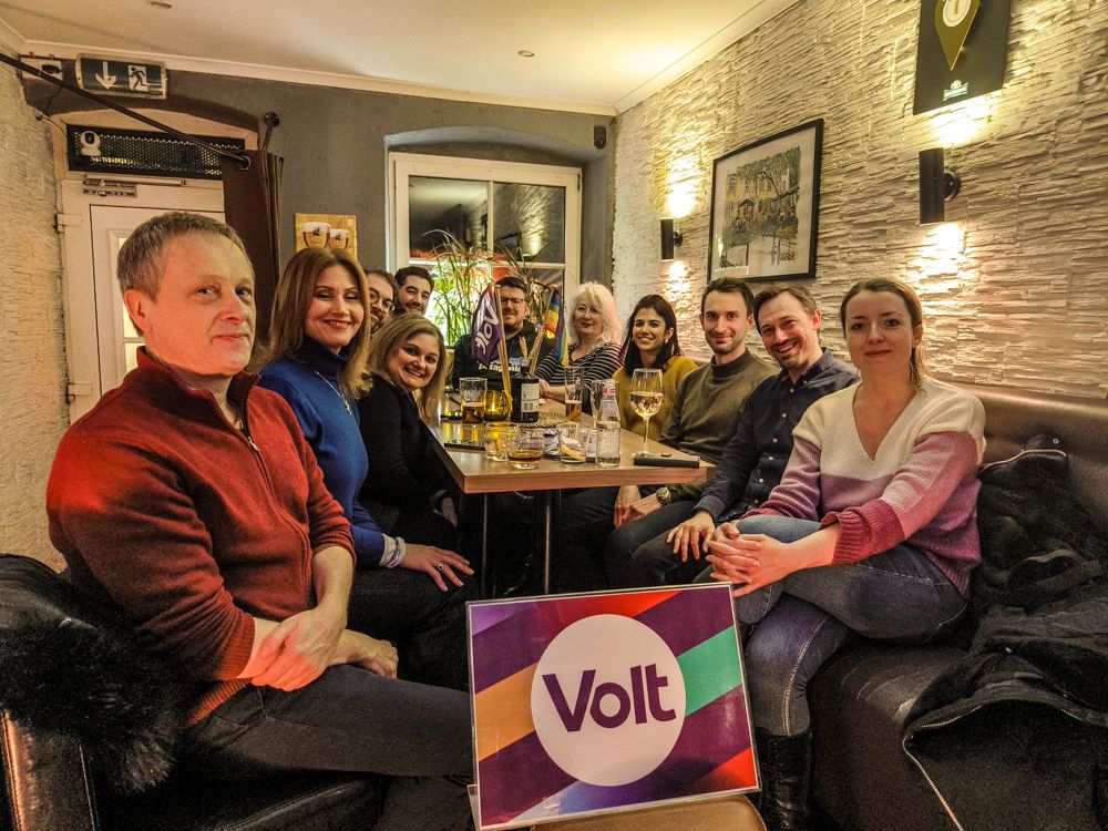 Group of people sittibg in a pub looking at the camera. A panel with a Volt logo is in the foreground. 