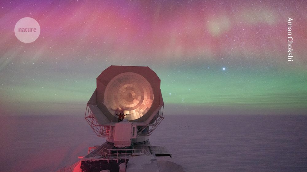 Two small figures pose for a photo on top of a large telescope dish which reflects the colours of the aurora in the sky over an Antarctic landscape