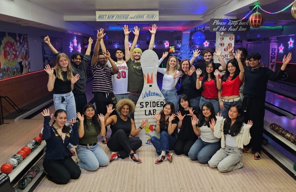 Group of people with arms in the air, smiling and cheering while in a bowling alley. A large sign shaped like a bowling pin is in the center of the group with black letters reading Spider Lab.