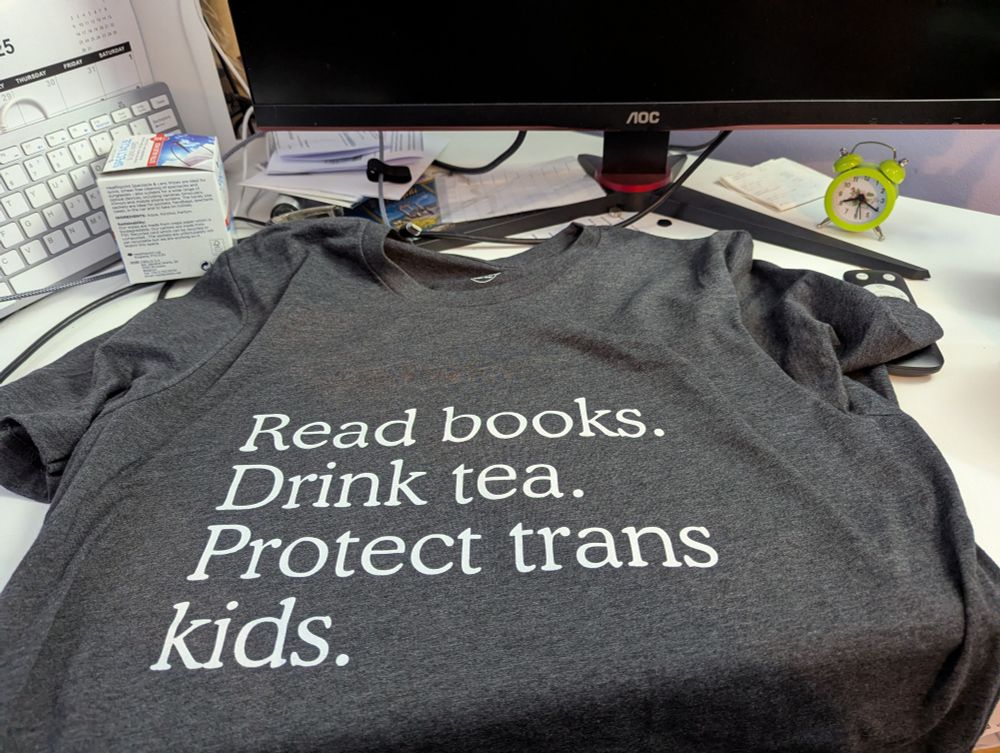 A grey t-shirt spread out on a desk. The writing on the t-shirt reads: Read books. Drink tea. Protect trans kids. 