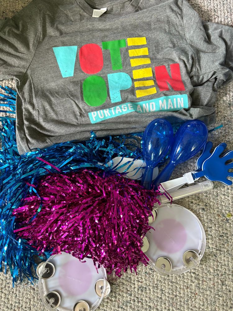 A "Vote Open Portage and Main"  grey tshirt surrounded by pink and blue party items - mini tamborines, shakers and shiny pom poms