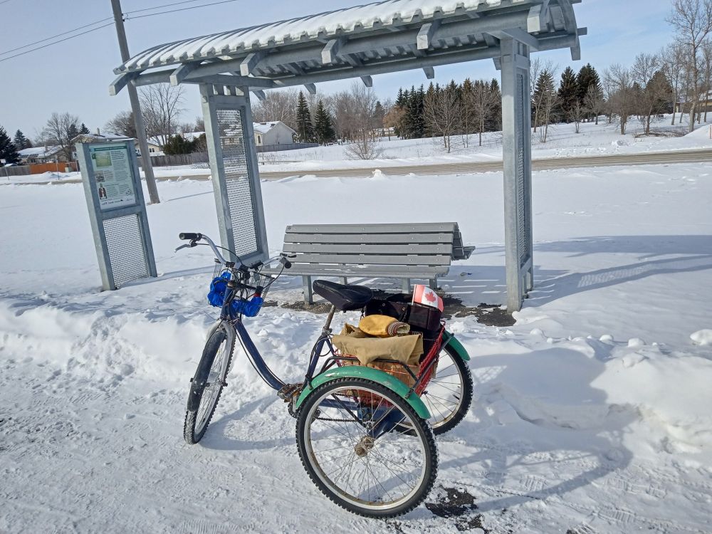 A cute trike with bright green fenders & a small Canada Flag parked in front of a bench shelter on a cold & sunny afternoon 