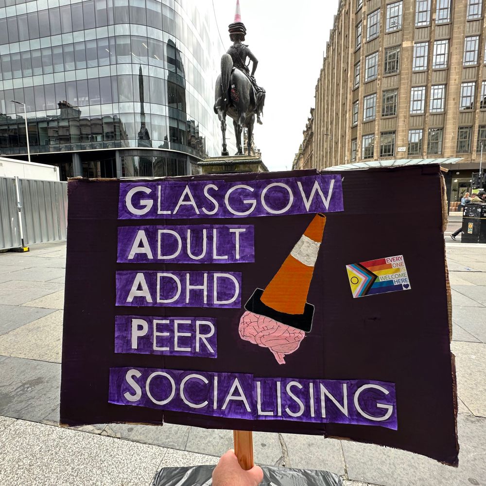 A photo of a hand held sign with our name and logo — a traffic cone hat on a brain, on a purple background 