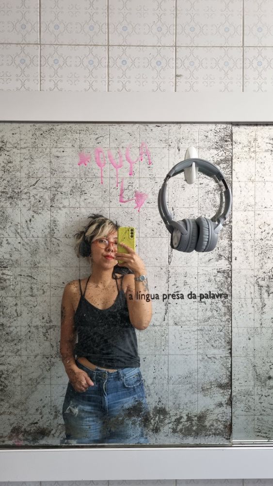 uma foto minha no espelho, sou uma mulher cis amarela, com uma mecha descolorida no cabelo, usando uma regata preta e uma calça jeans. no espelho tem o escrito "a língua presa da palavra" 