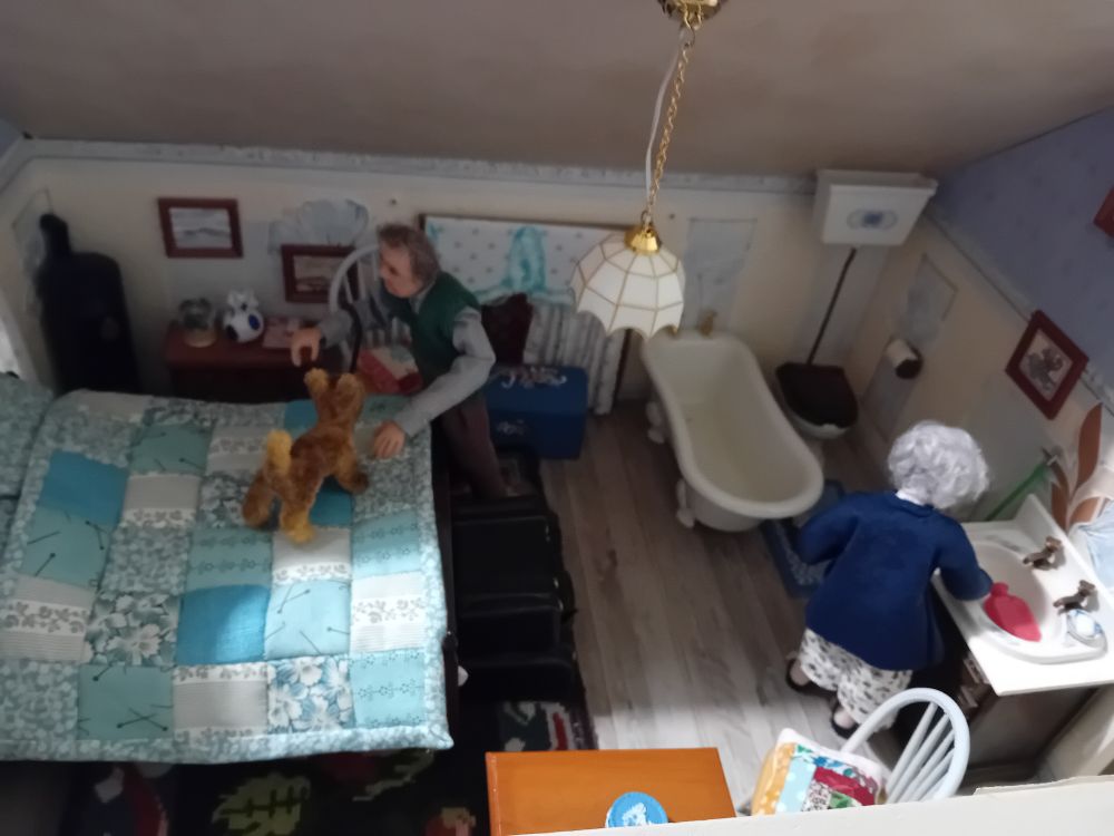 A dolls house interior. In the bedroom an elderly lady fills a hot water bottle at the sink, while her husband pets their dog who is standing on the bed.