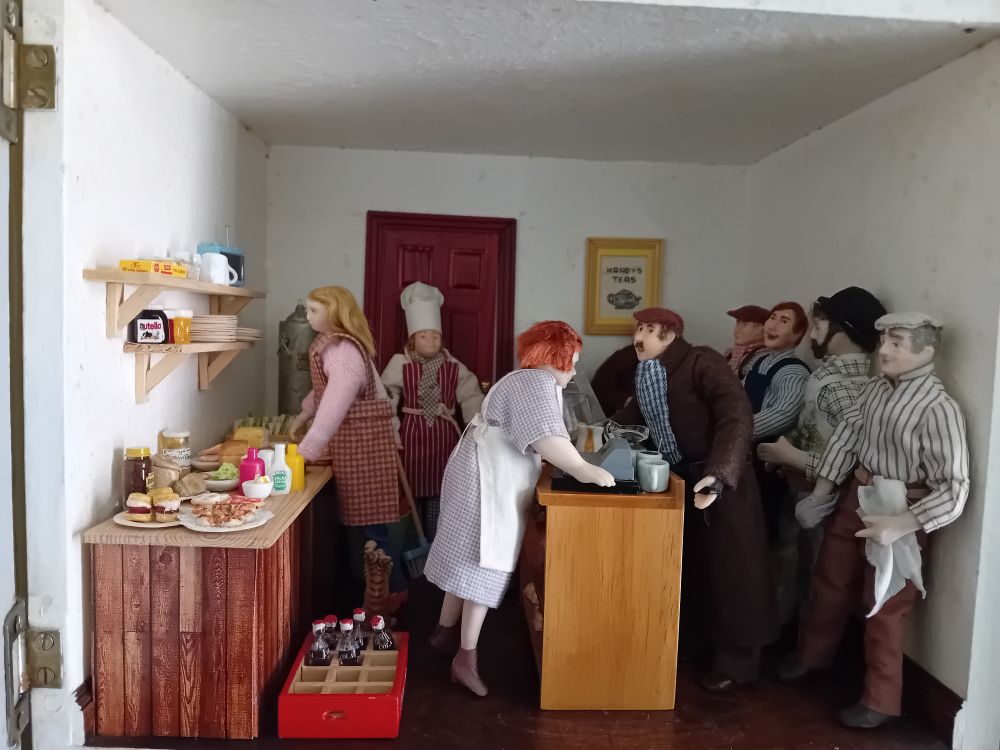 A dollshouse scene: a crowd of workmen queue in a small shop while people make tea and sandwiches.