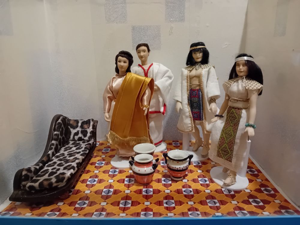 Four dollshouse figures stand in a room. One couple are classical Romans, the other couple are ancient Egyptians. Some urns stand on a floor that looks more like 1950s lino. A low couch is covered in leopard print. The overall effect is underwhelming
