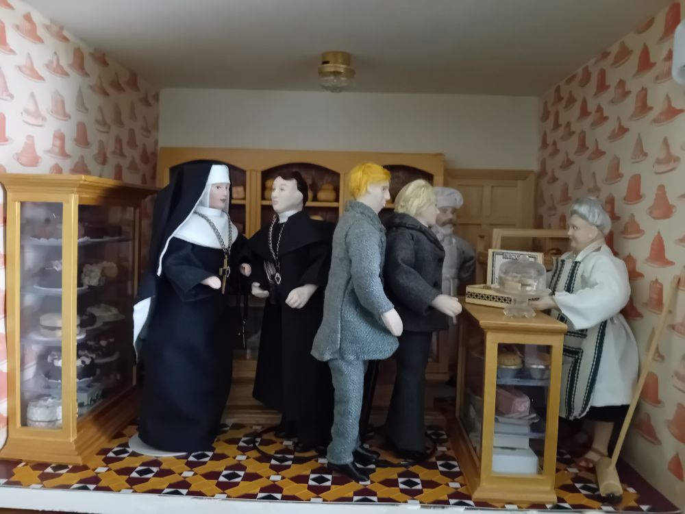 A dollshouse scene: vicars, a priest and a nun wait to be served in a bakery.