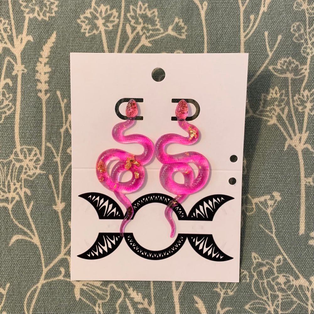 Pink resin snake earrings with gold foil inclusions on a white earring card. Green and beige floral cloth background