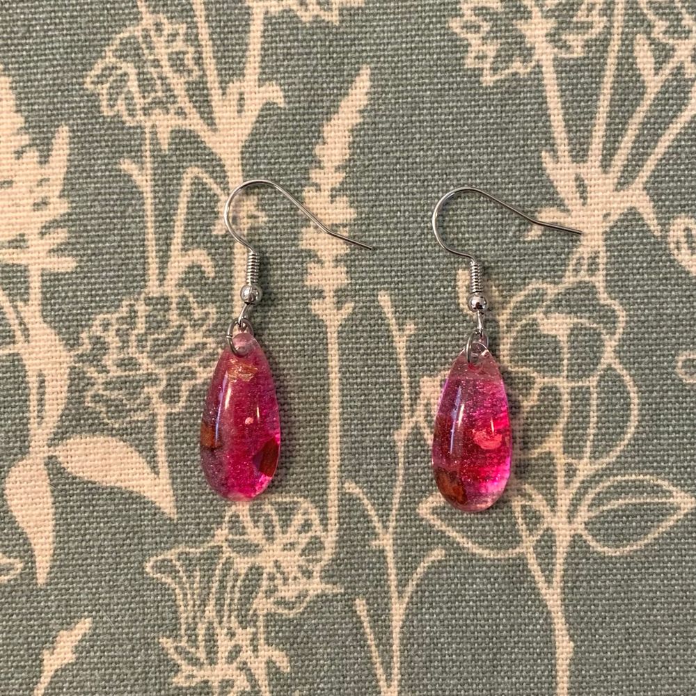 Pink resin teardrop earrings with gold foil and rose petal inclusions on a green and beige floral cloth background 