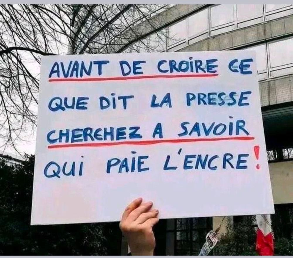 Une main tenant une pancarte avec écrit dessus "avant de croire ce que dit la presse, cherchez à savoir qui paie l'encre!"
