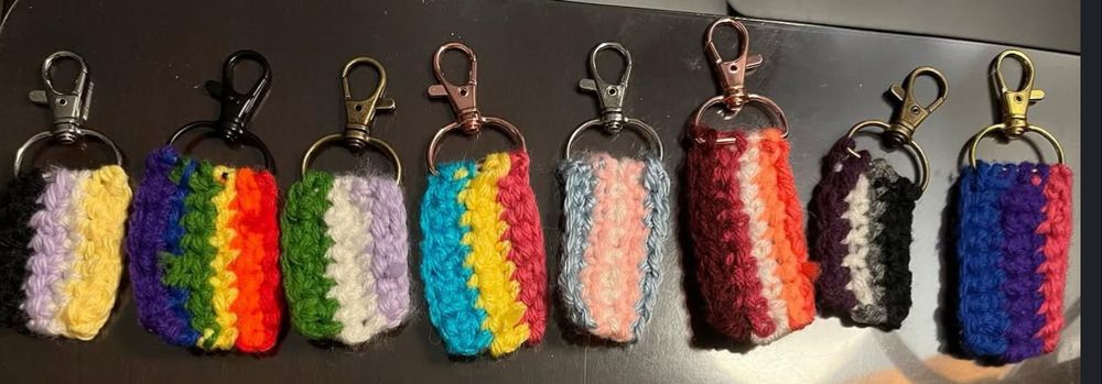 8 crochet pride flag keychains in a row. from left to right: nonbinary flag (yellow, white, purple, black), rainbow flag (red, orange, yellow, green, blue, purple), genderqueer flag (purple, white, green), pansexual flag (pink, yellow, blue), transgender flag (light blue, pink, white, pink, blue), lesbian flag (dark orange, light orange, white, pink, maroon), asexual flag (black, grey, white, purple), bisexual flag (pink, purple, blue).