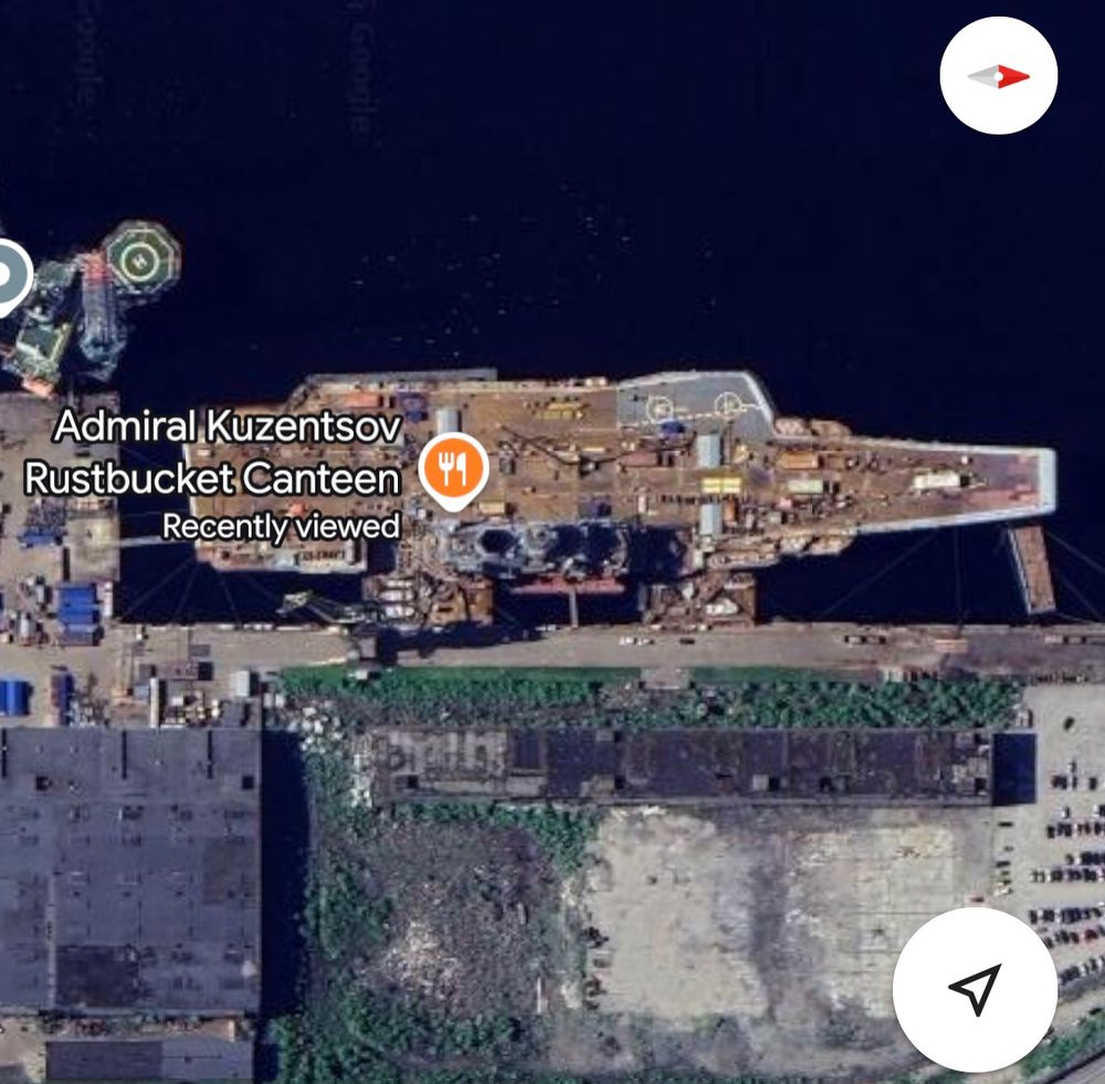 A satellite view showing the Admiral Kuznetsov aircraft carrier docked at a shipyard. The vessel is visibly rusted and under maintenance. A food icon and label mark the ship as the location of the “Admiral Kuzentsov Rustbucket Canteen"