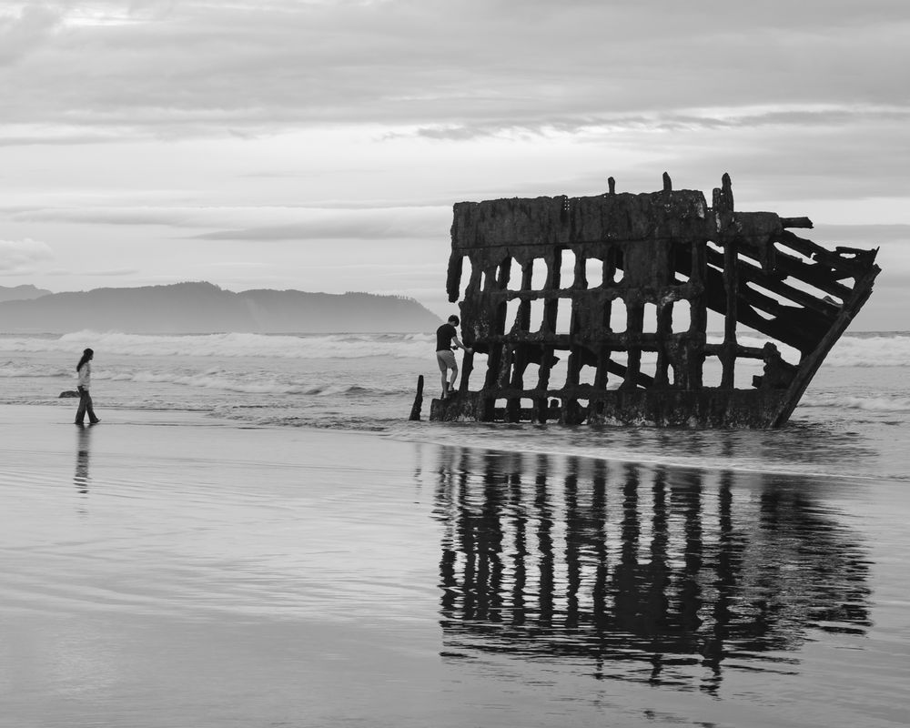 A black and white image of the rusted metal remains of a shipwreck with one kid climbing up the front and another walking toward it. The remains of the wreck are small and don't particularly appear ship-like.