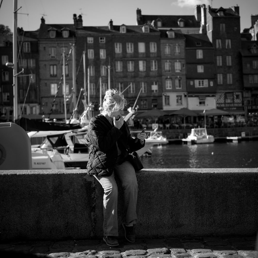 Woman sitting on the harbour wall puzzling over her phone with boats and buildings beyond.