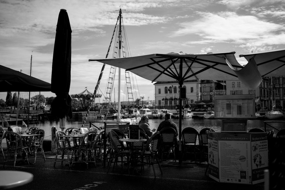 Harbour scene: tables, chairs, and big umbrellas. Boats and buildings beyond as a coupe sit at a table and enjoy the scene.