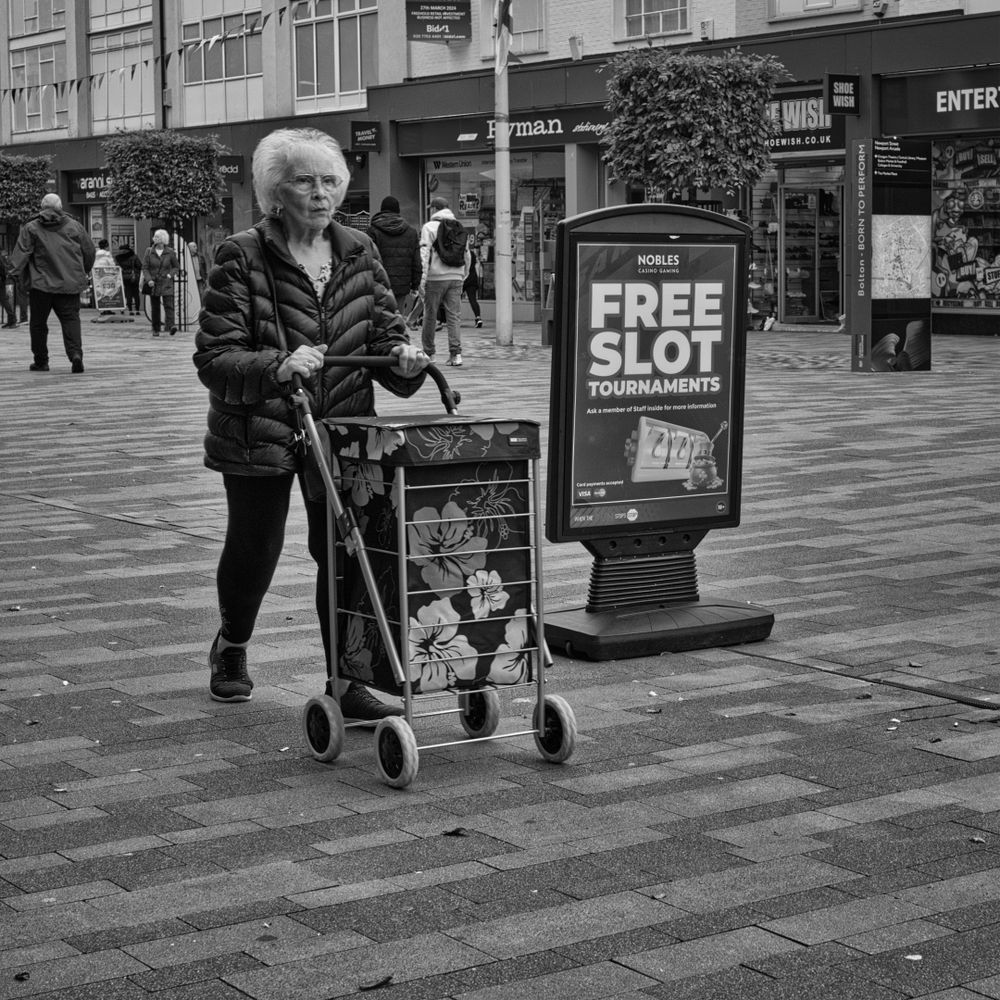 Woman pushing her shopping trolley past an advert for the local slot machines. Shops form the background to a pedestrian area.