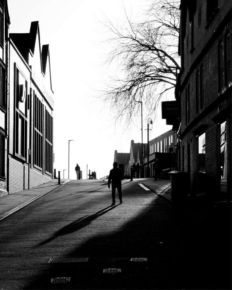 Office buildings on both sides of a narrow street cast strong shadows from one side. Figures in the distance with a lone figure in the mid-ground casting a strong shadow