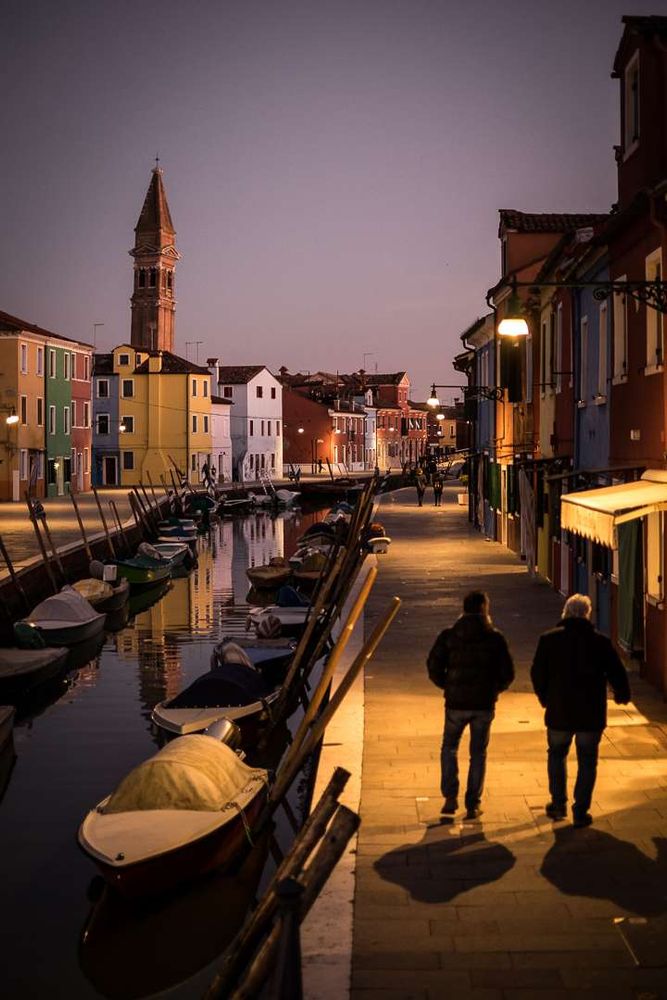 Two men walking along a canal-side pavement after dark in Burano, Venice