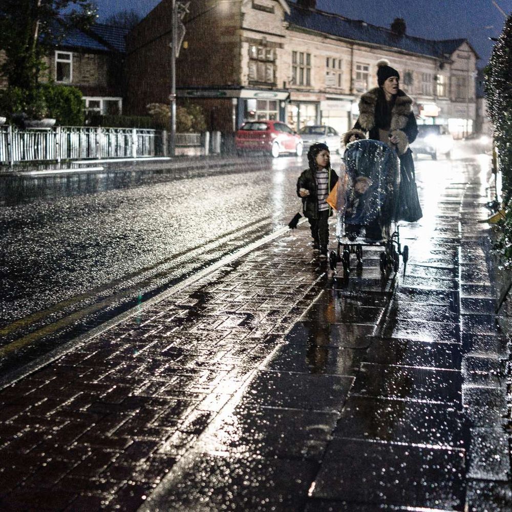 Mother and child with another in pushchair walking on a rainy night, illuminated by car headlights