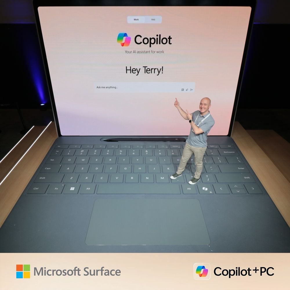A man in a gray Microsoft polo shirt is standing on a giant Surface keyboard, pointing enthusiastically at a large screen that displays ‘Copilot: Your AI assistant for work’ and the message ‘Hey Terry!’ The Microsoft Surface and Copilot+PC logos are visible at the bottom.
