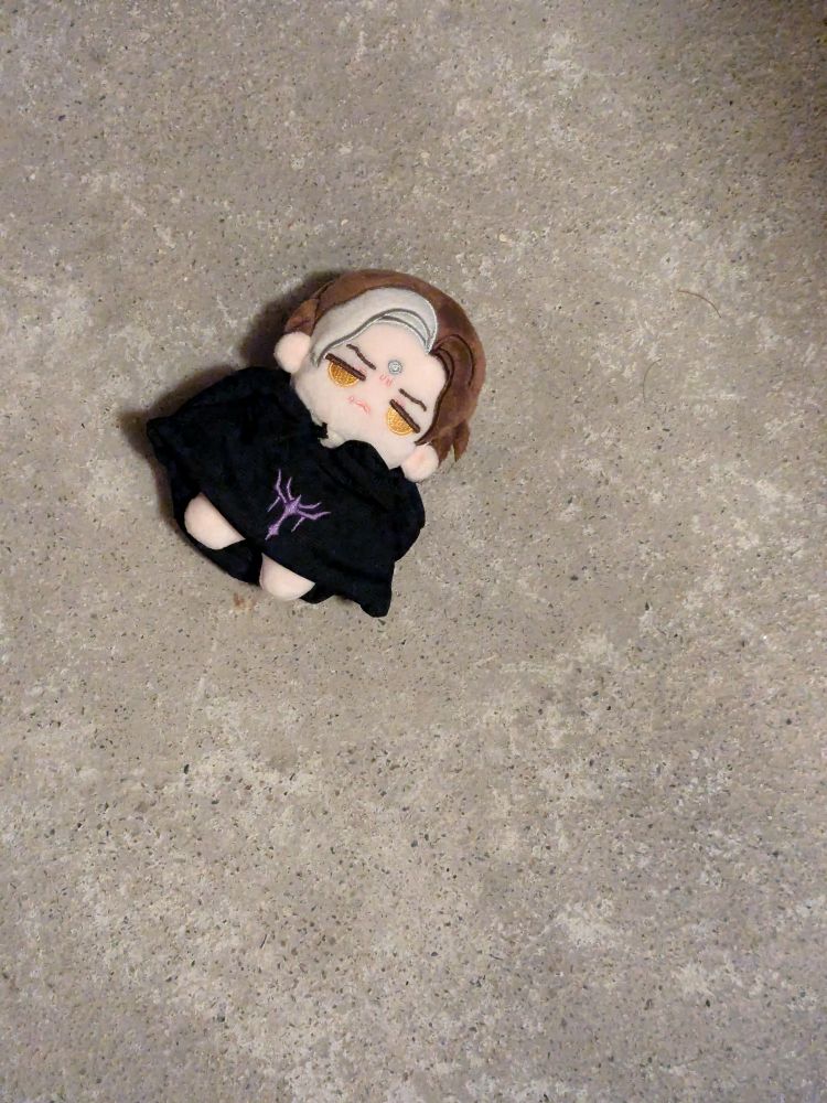 a small plush doll of Final Fantasy 14 character Emet-Selch lying on a beige-grey concrete floor.